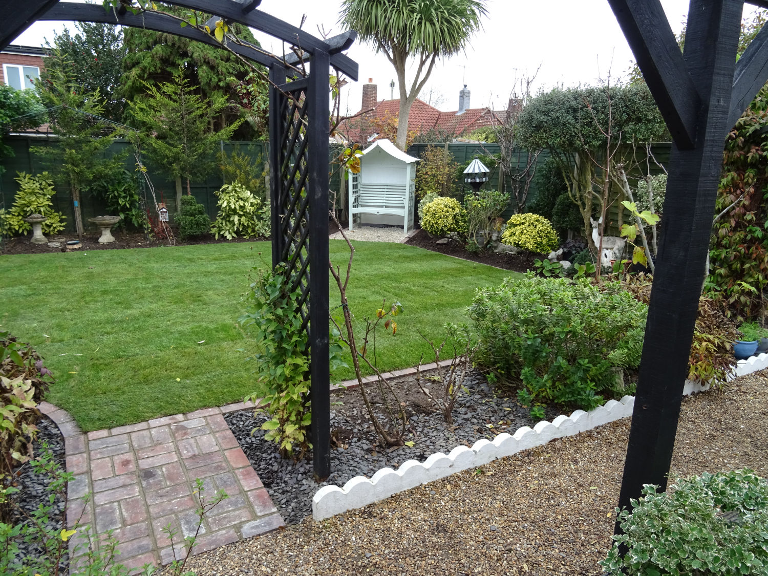 Rear garden with gravel pathway leading through a pergola onto the lawn surrounded by borders of shrubs and a garden seat